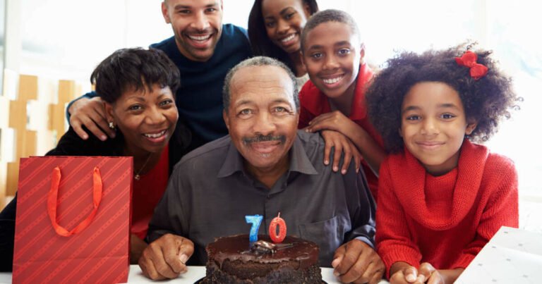 Man sitting down with a birthday cake celebrating his 70th birthday with his family stood next to him.