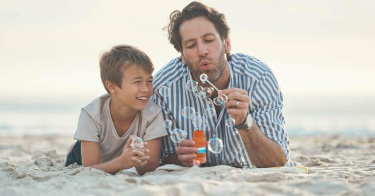 Playful father lying on the beach, blowing bubbles with his son.