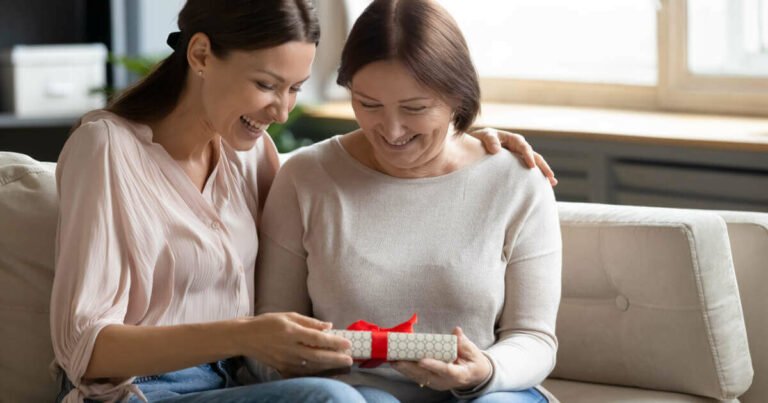 Capturing a heartwarming moment at home, a grown-up daughter sits beside her mother, offering a beautifully wrapped gift.