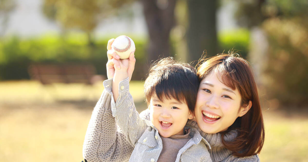Mother and son share a smile while sitting together, holding a baseball in a field.