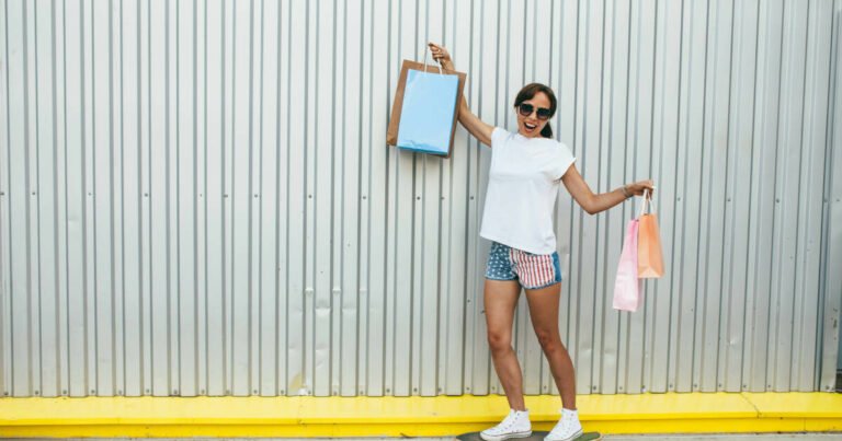 Woman confidently rides a skateboard while doing shopping, holding up her shopping bags and smiling.