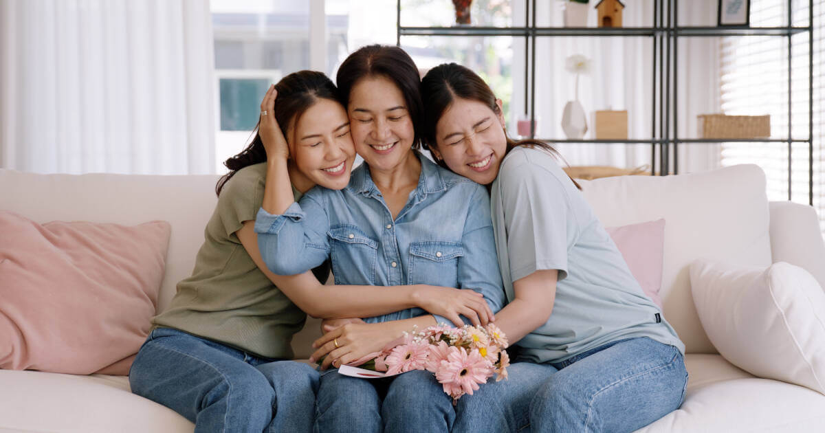 Mom sits on sofa, embraced by two smiling daughters with flowers on her lap.