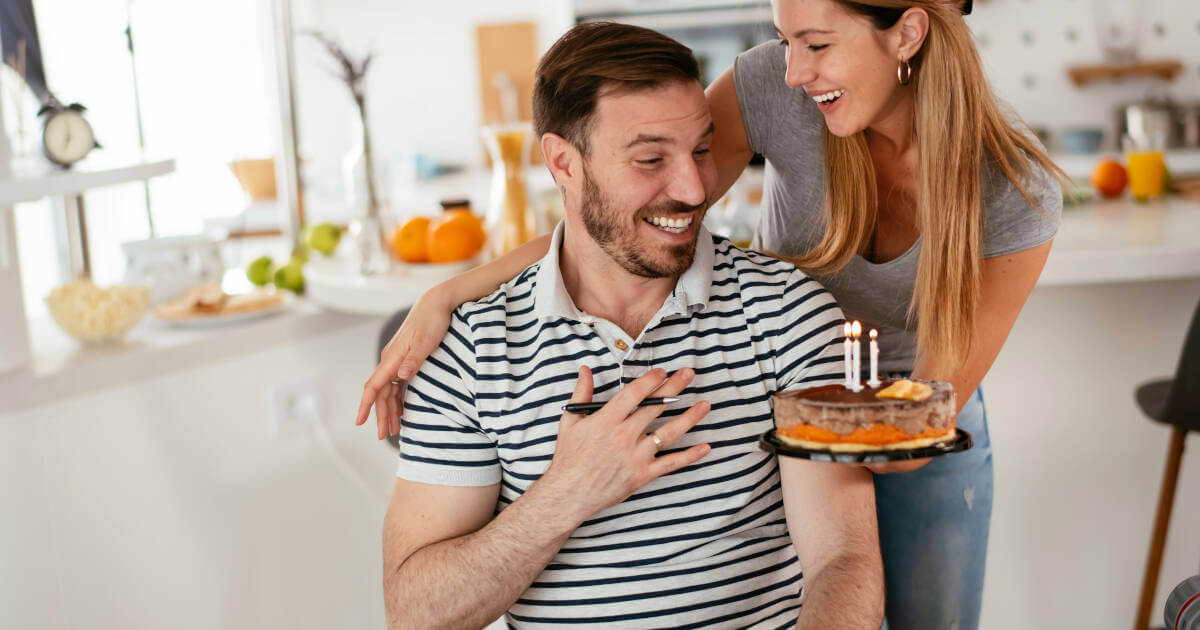 Woman surprises her sitting boyfriend with a birthday cake with candles in their kitchen on his special day.