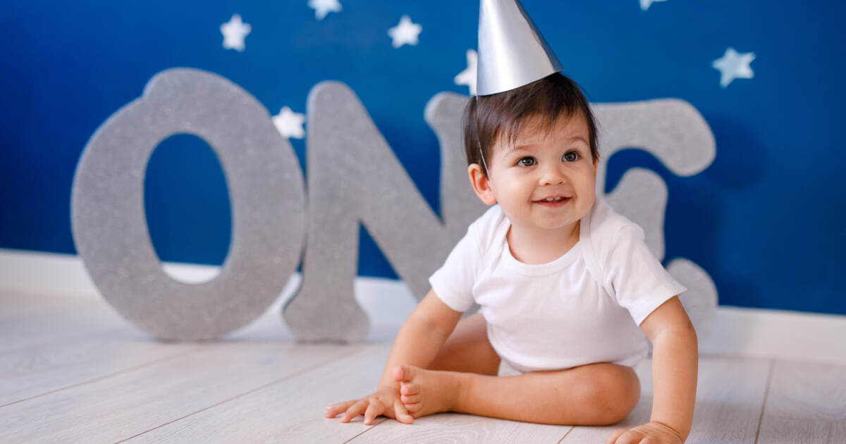 1 year old baby boy, donning a white t-shirt and a silver hat, surrounded by silver letters spelling 'ONE' on a purple background.