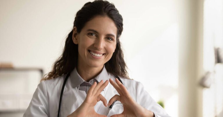 Smiling female GP in medical uniform shows heart gesture.