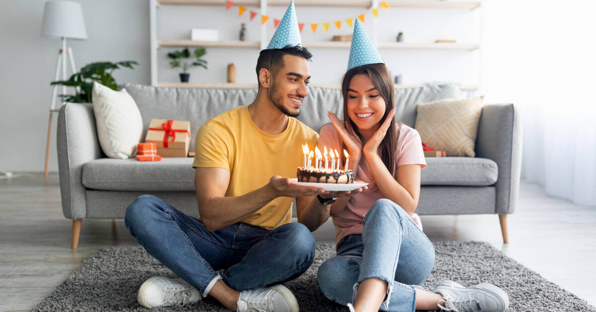 Happy millennial couple wearing party hats, joyfully celebrating the wife's birthday with a festive cake at home.