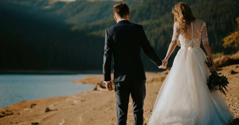 Bride and groom eloping in their wedding gowns, holding hands and hugging in a serene desert-like landscape by the lake.