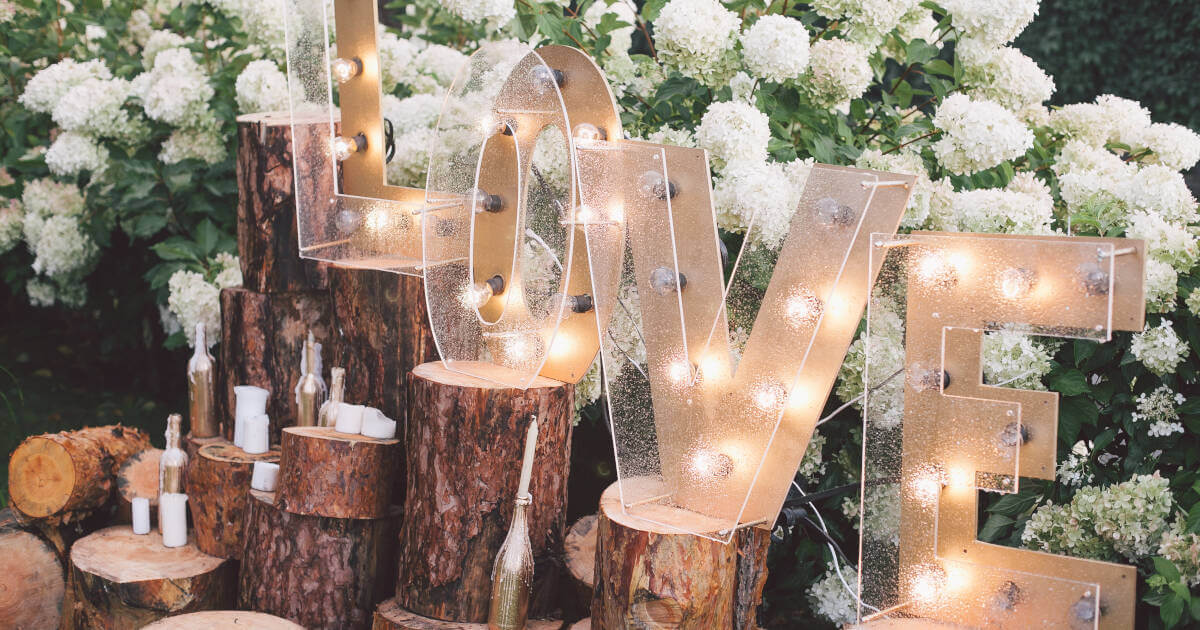 The word 'Love' beautifully lit up on logs at an enchanting engagement party, flowers in the background.