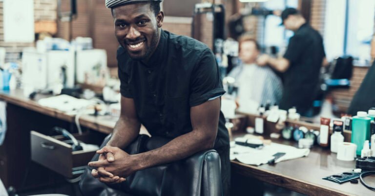 The barber stands confidently behind his chair, looking at the camera, against the backdrop of a busy barbershop.