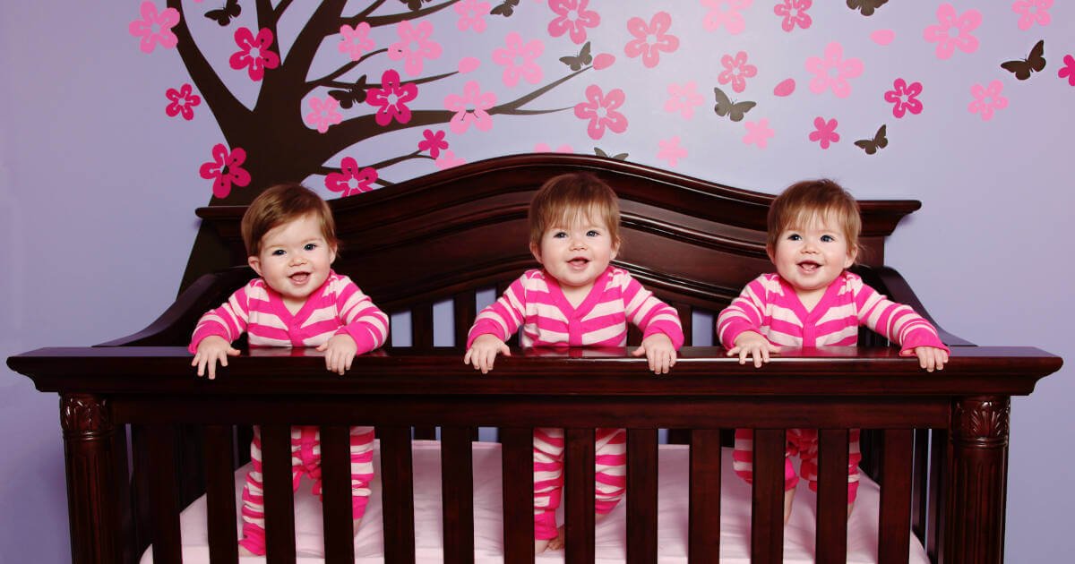 Triplets in matching pink outfits, standing in cribs, all smiles. Pink wallpaper with a tree and butterflies in the background.