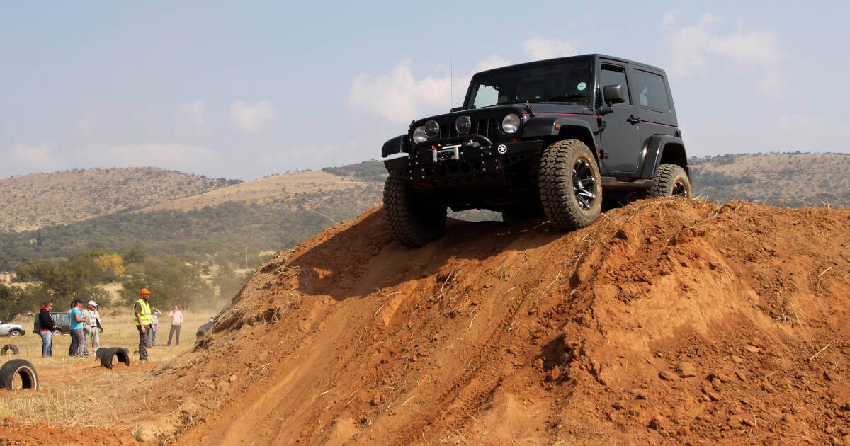 Black Jeep Wrangler parked atop a sand mound, with visible tire tracks and people in the background.