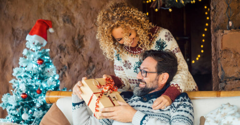 A happy couple enjoys the holiday season at home, sharing presents. The woman surprises her husband by giving him a Christmas gift from behind.