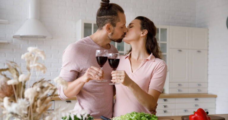 A happy, loving couple kissing and clinking glasses of red wine, celebrating their wedding anniversary or enjoying a romantic home date in a modern kitchen, expressing their affectionate feelings.