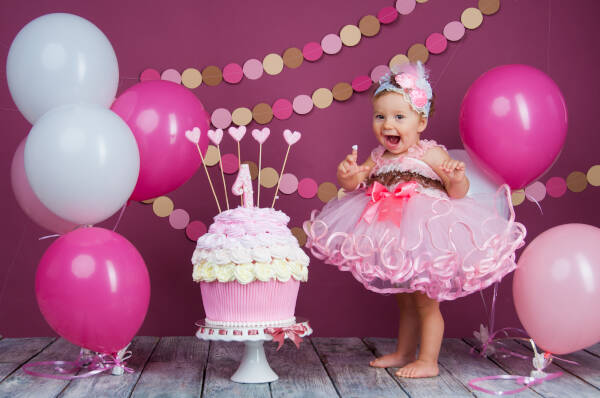 1-year-old girl squeals with joy next to a pink cake surrounded by pink and white balloons against a pink backdrop