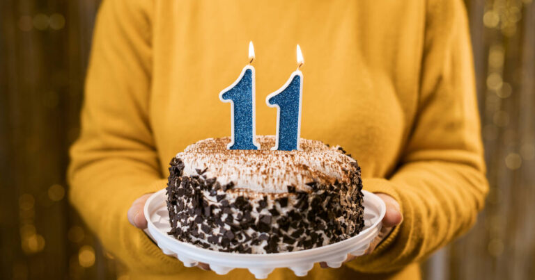 A woman holding a birthday cake with burning number 11 candles against a decorated background.