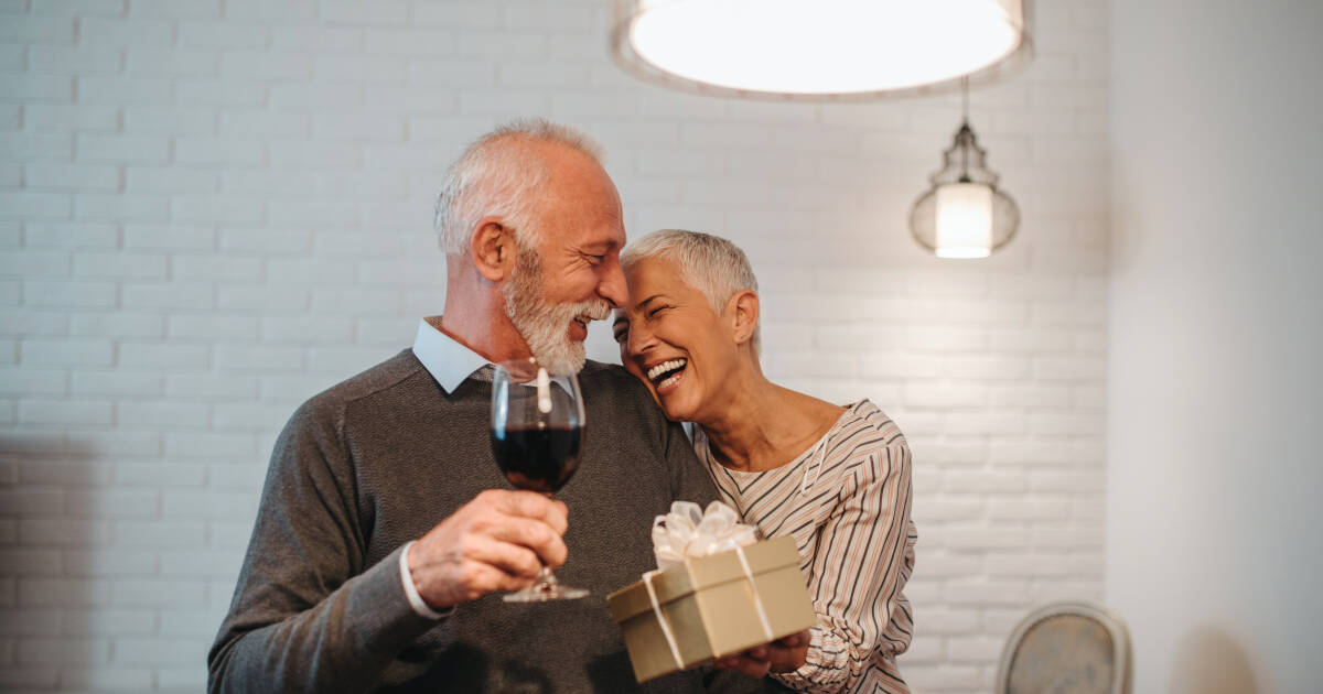 Husband enjoys a glass of wine as wife surprises him with a present, and they share laughter together.