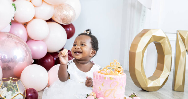 Laughing 1-year-old girl celebrates her first birthday with a cake to smash and balloons in the background.
