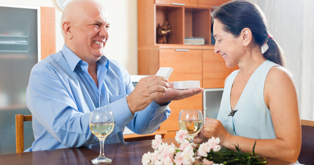 A mature man presenting a woman with a jewelry box at a table.