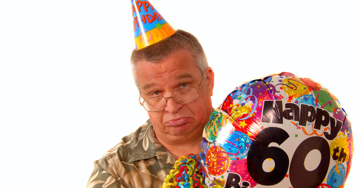 Man wears a party hat with a sad expression, holding a balloon for his 60th birthday celebration.