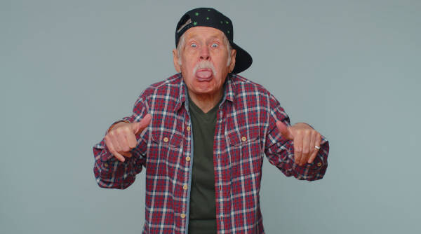 80-year-old man makes playful faces, tongue out, wearing a tilted baseball cap, embracing youthful spirit