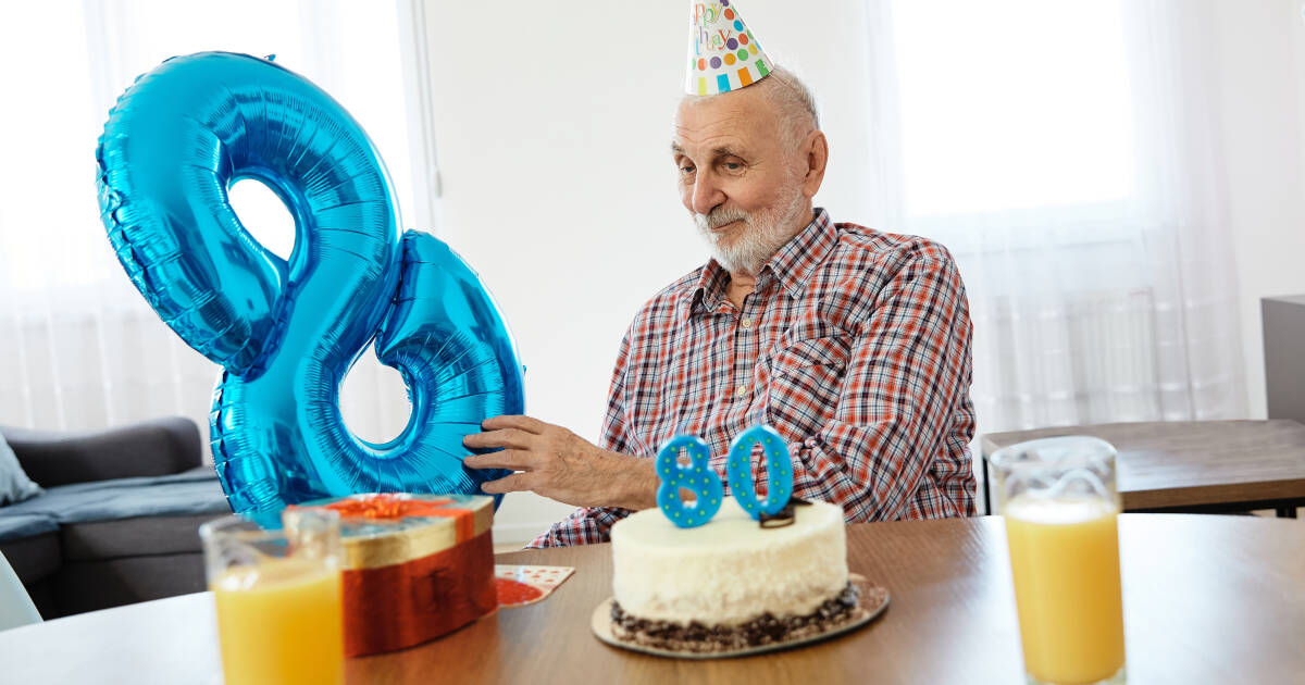 Elderly man, wearing a party hat, celebrating his 80th birthday alone at home with cake.