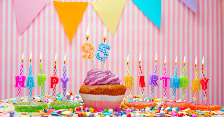 Against a pink background, a birhday celebration table with muffins topped with pink cream and lit happy 95 birthday candles.