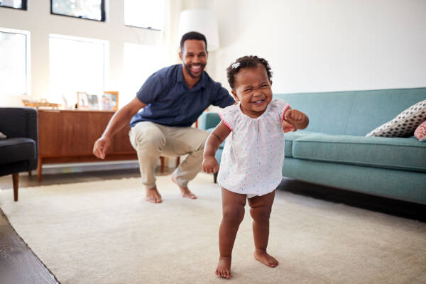 A baby girl dances in the living room while her uncle looks on, crouched down and smiling.