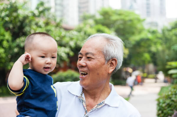 A grandfather holds his baby grandson as they stand together outside in the park.