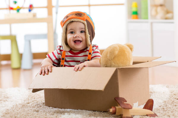 A happy pilot aviator baby boy with a teddy bear toy plays in a cardboard box.