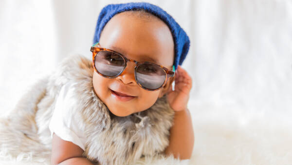 A hipster baby wearing a fur vest and sunglasses lies on a white bed in a room with curtains