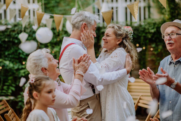 A mature couple embracing each other, celebrating their wedding with family cheering in a backyard setting.