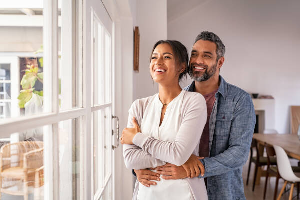 A middle-aged couple stands together by a window in their house, smiling as the man hugs the woman from behind.