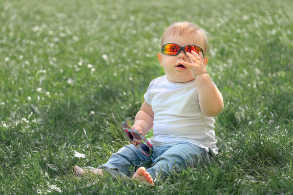 A one-year-old boy sits on a green lawn wearing sunglasses on a summer day