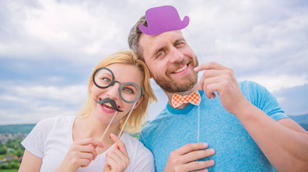 A smiling man and woman holding silly props, woman holds up a moustache and glasses, the man holds a small purple hat cardboard prop.