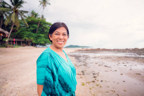 A woman stands on a sandy beach, smiling widely at the camera, with palm trees in the background.