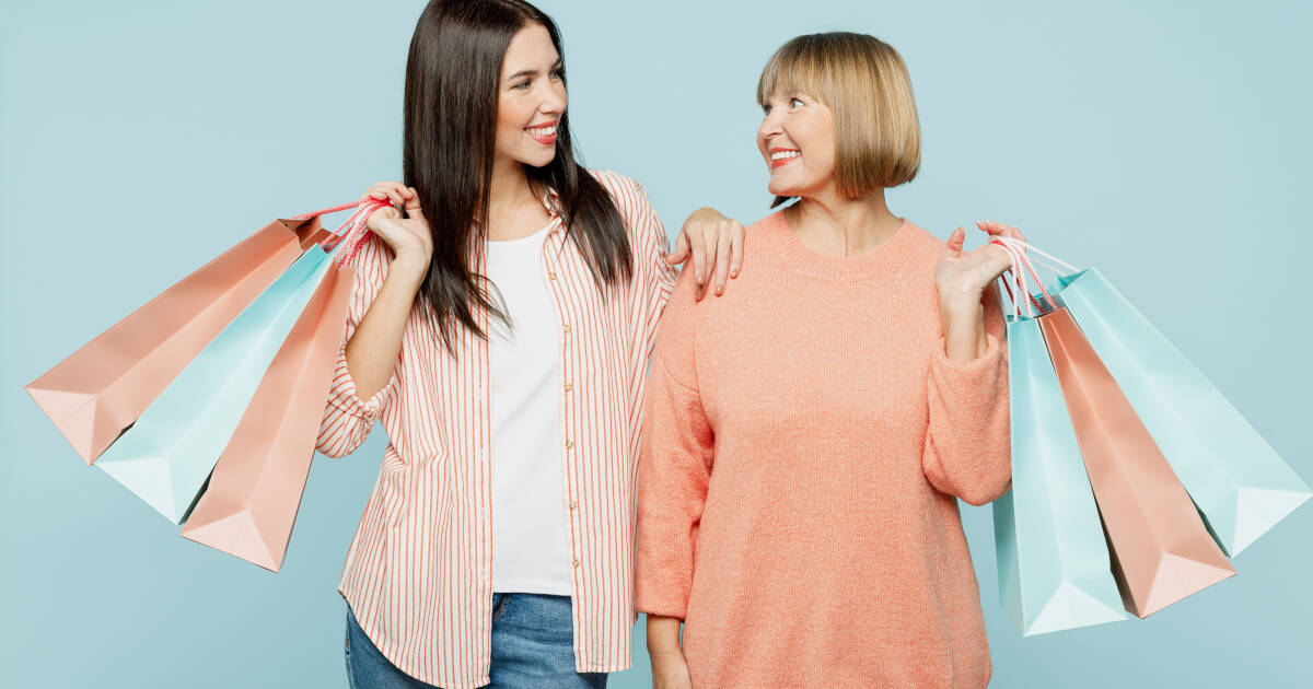 Elderly mom and young adult daughter, both wearing casual clothes, hold gift bags and smile on light blue background