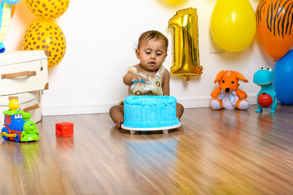 Baby boy celebrates his first birthday with a blue cake and '1' balloon amidst toys on wooden floor
