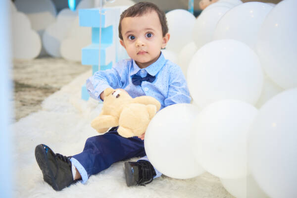 Baby boy sits on the floor amidst a pile of white balloons, holding a teddy bear and staring at the camera.