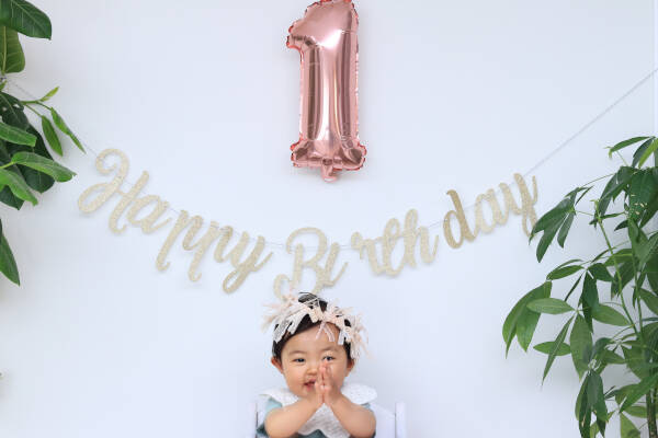 Baby girl sits smiling with prayer hands, Happy Birthday sign and '1' balloon in background on white backdrop