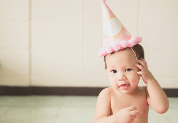 Baby girl smiles wearing a pink party hat at home, with one hand on her head