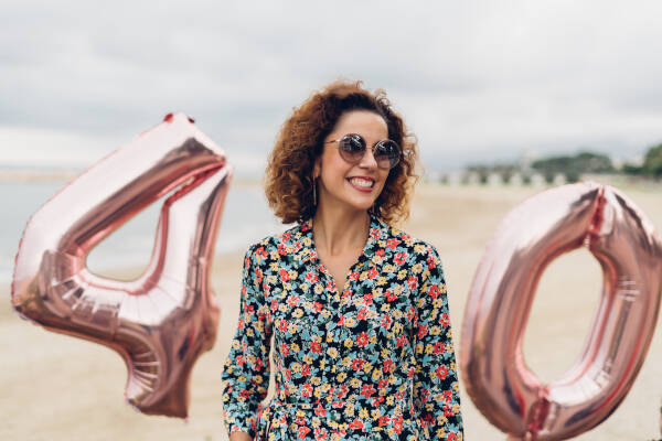 Beautiful curly brunette celebrating her 40th birthday with large balloons, walking along a beach.