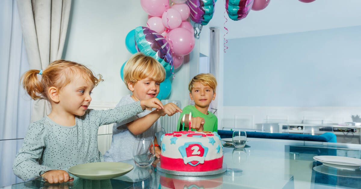 Three joyful children at a birthday party excitedly point at the cake for a 2-year-old's celebration.