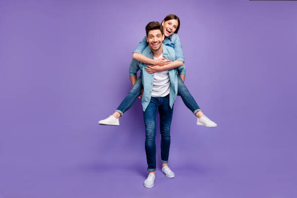 Boyfriend gives his girlfriend a piggyback ride as they both smile in front of a purple backdrop