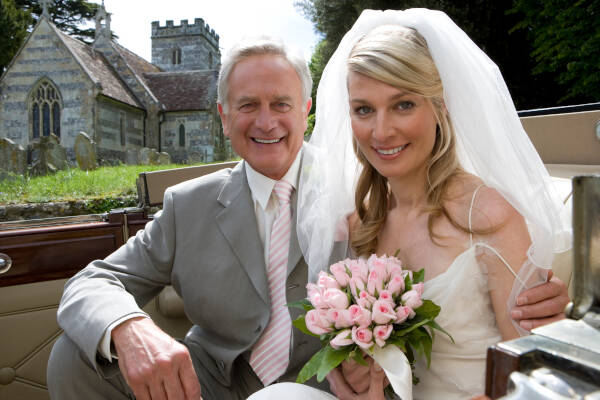 Bride and grandfather smile at the camera in a vintage car in front of a church, in a horizontal waist-up portrait.