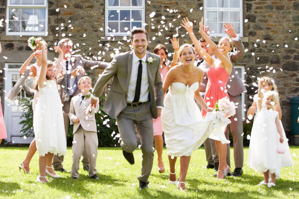 Bride and groom running towards the camera outdoors with massive smiles, guests throw confetti behind them