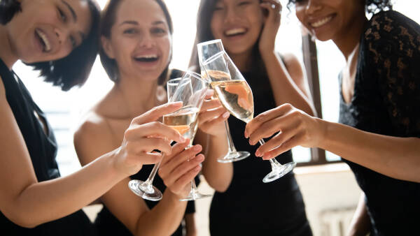 Close-up focus on glasses of champagne joined together, with four women smiling and celebrating an engagement.