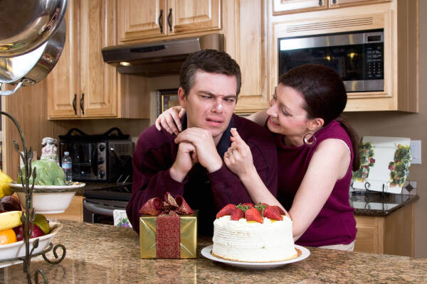 Couple horsing around in kitchen with gifts and cake. He makes faces, trying to stare at frosting she put on his nose