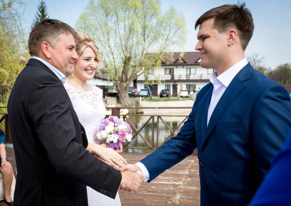 Father shakes his son's hand on his wedding day, bride standing next to groom's father, smiling