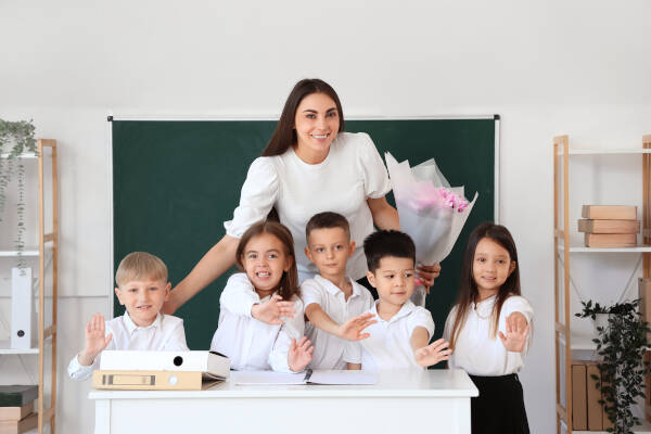 Female teacher holds flowers, posing for photo with little school children in classroom