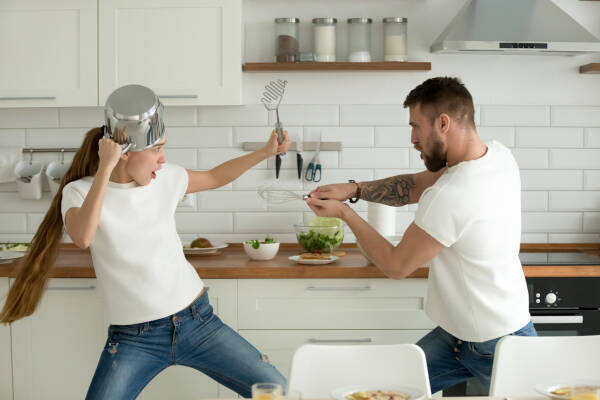 Funny couple playfully pretending to fight with utensils and tools while cooking together at home.